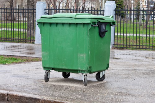 A large green plastic wheelie bin with a closed lid, positioned on a concrete pavement next to a curb, with four black wheels clearly visible at the base. The bin's surface appears slightly textured and weathered, indicating regular outdoor use. In the background, there is a black metal fence with vertical bars and decorative posts, behind which is a grassy area with leafless trees and a pathway, suggesting an urban or suburban setting. The scene is evenly lit, reflecting a cloudy day, and the environment appears clean with no visible rubbish inside or outside the bin. This image illustrates typical rubbish collection equipment used by waste management services such as Office Clearance Temple servicing areas in the vicinity of Temple and nearby postal districts, emphasizing the practical aspect of waste disposal and rubbish removal in residential or commercial contexts.