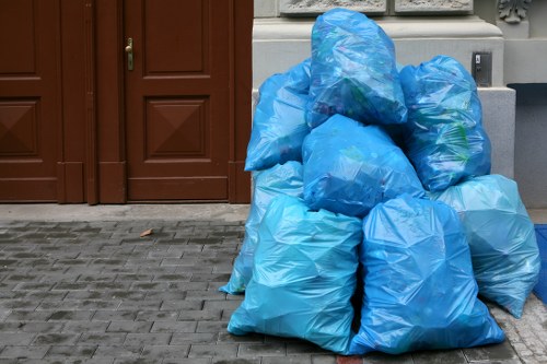 A collection of large blue plastic rubbish bags filled with waste material, stacked outside a building on a paved sidewalk area. The bags are made of thin, slightly glossy plastic and are securely tied at the top. They are positioned close to a brown wooden door with panel details, and are situated on a grey stone or concrete pavement. In the background, part of an ornate grey metal fence and a white wall are visible, set against an urban environment typical of a residential or commercial property in a town. The scene appears to be part of a rubbish removal or waste collection effort, demonstrating the typical waste packaging used in office or property clearance services such as those provided by Office Clearance Temple in the local area.