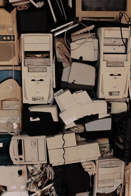 A collection of black rubbish bags and scattered discarded materials, including cardboard and plastic, piled up in front of a red metal door set into a beige, textured wall. The door features multiple