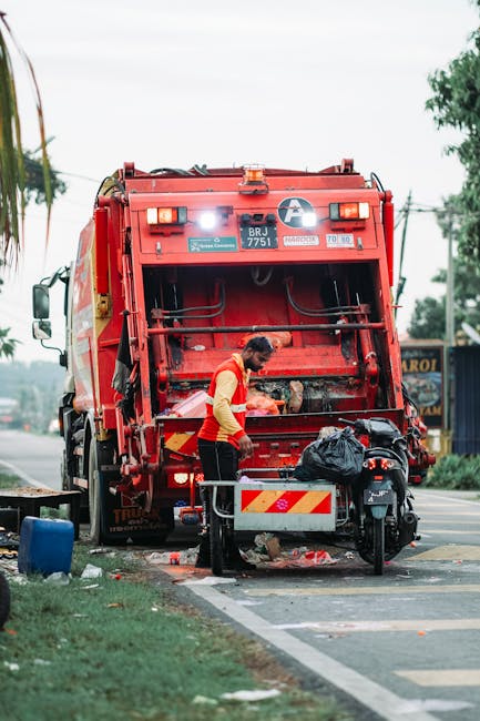 Two waste management workers are engaged in rubbish removal outdoors in an urban setting, standing next to a yellow skip loader on a paved surface. The worker in the foreground, wearing a teal uniform