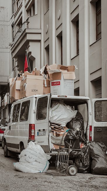 A white commercial van parked on a city street with its rear doors open, revealing a large load of mixed rubbish and waste materials. The cargo area is filled with various items including black trash
